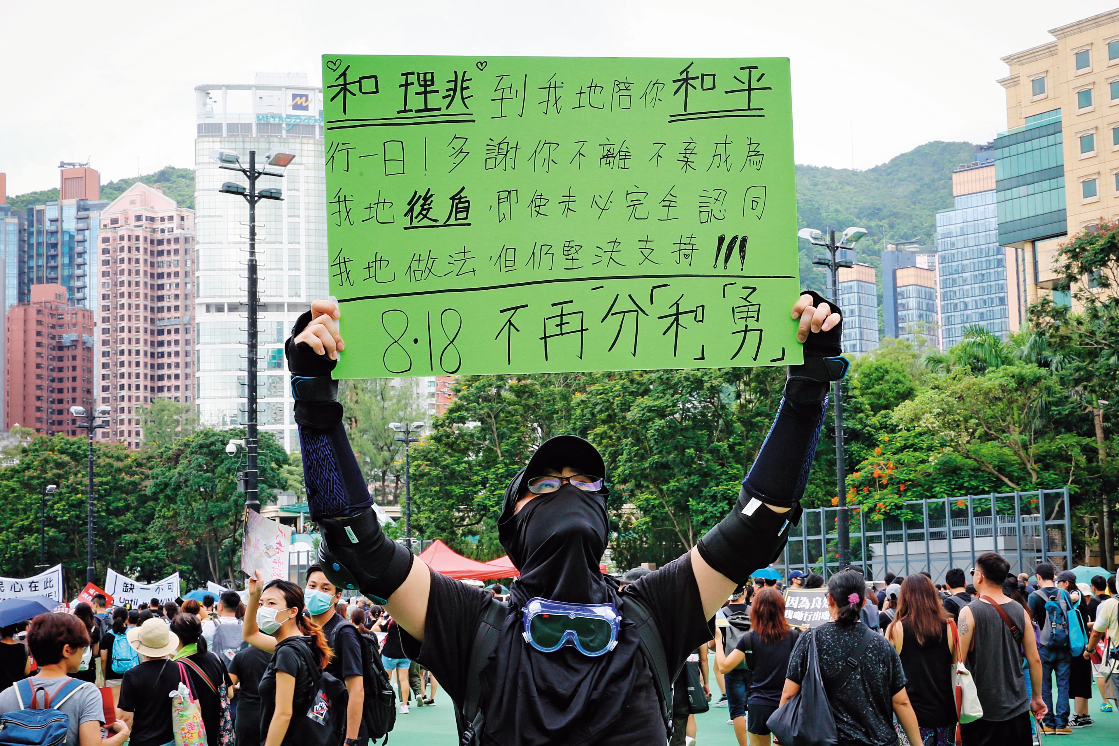威爾(化名)高舉自備手寫看板,主張「818不再分『和』『勇』」,頂著高溫,在維園的一角苦站。