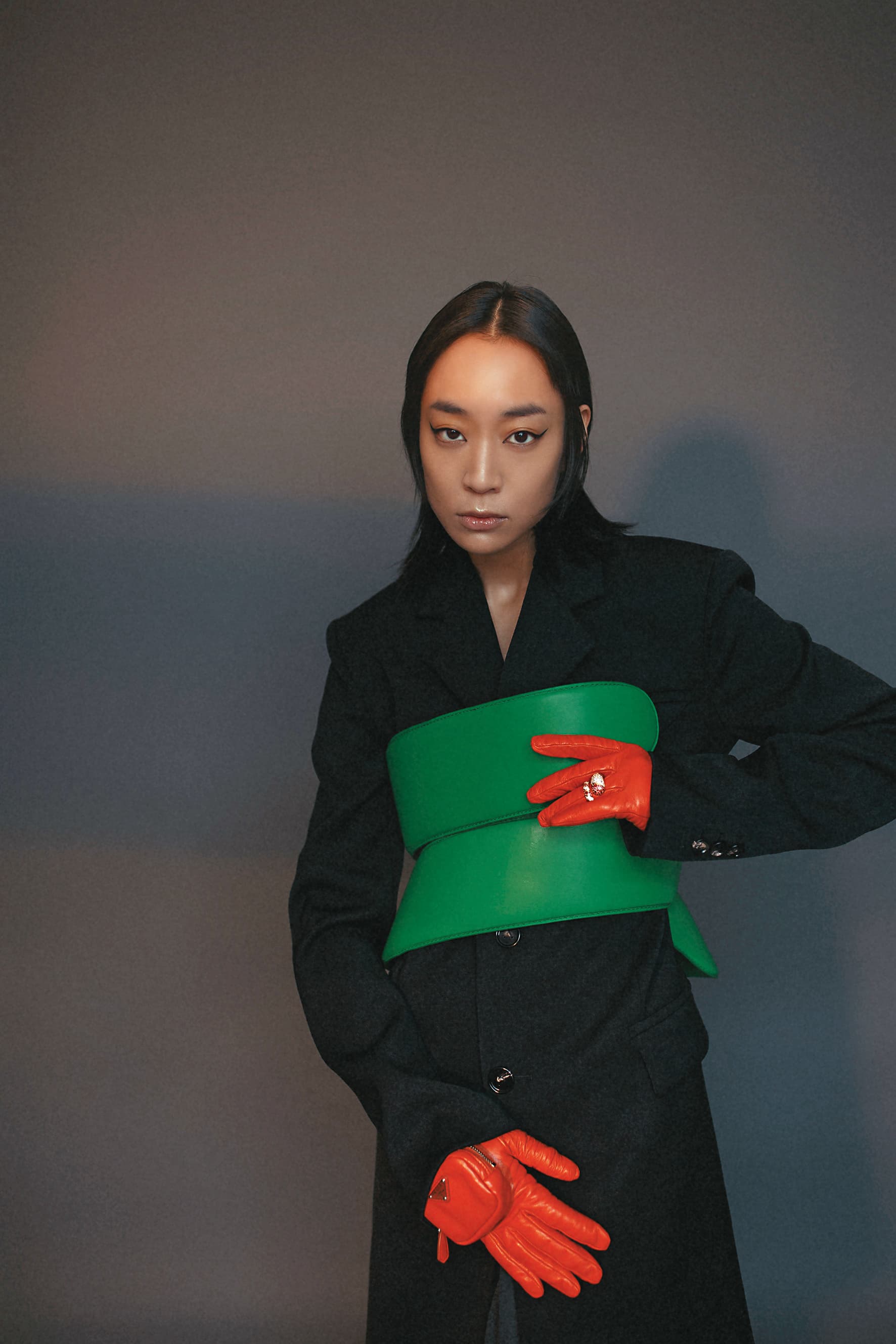 黑色單側雙口袋羊毛外套 NT$96,500 by BOTTEGA VENETA；鸚鵡綠螺旋繞身皮帶 NT$81,700 by BOTTEGA VENETA；紫紅白鋯石純銀鍍金戒指 NT$51,300 by BOTTEGA VENETA；橘色口袋皮革手套 NT$34,000 by PRADA。