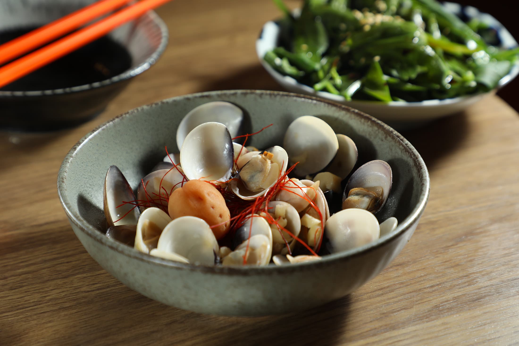 「梅酒蛤蠣」(前,200元/份)以自釀梅酒嗆炒,吃得到濃郁海味與酒香;「小農市集時蔬」(後,50元/份)可品嘗到在地季節蔬菜。
