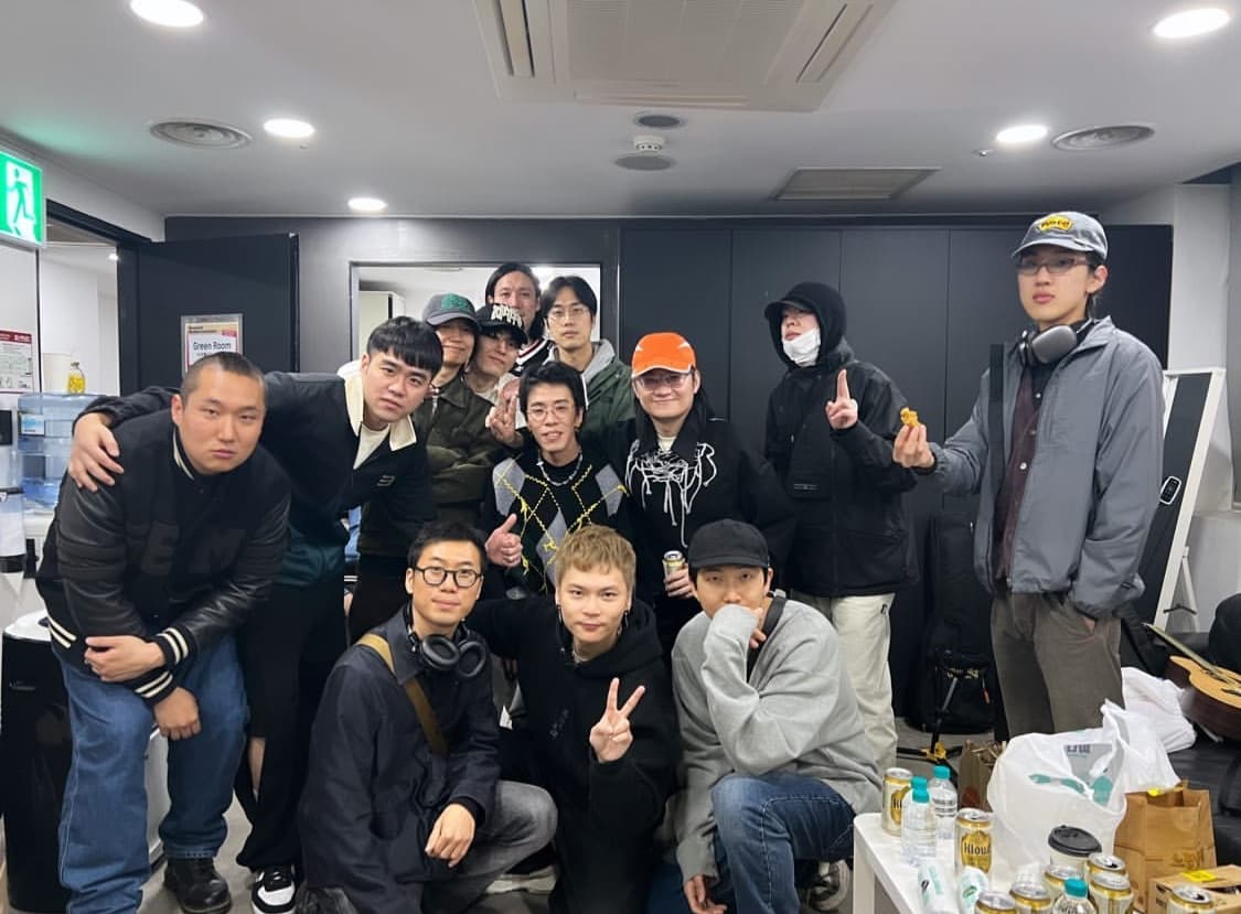RM(前排右)一身灰色衛衣牛仔褲搭配帽子的輕便裝扮,在落日飛車表演後台一起大合照。(翻攝落日飛車IG)